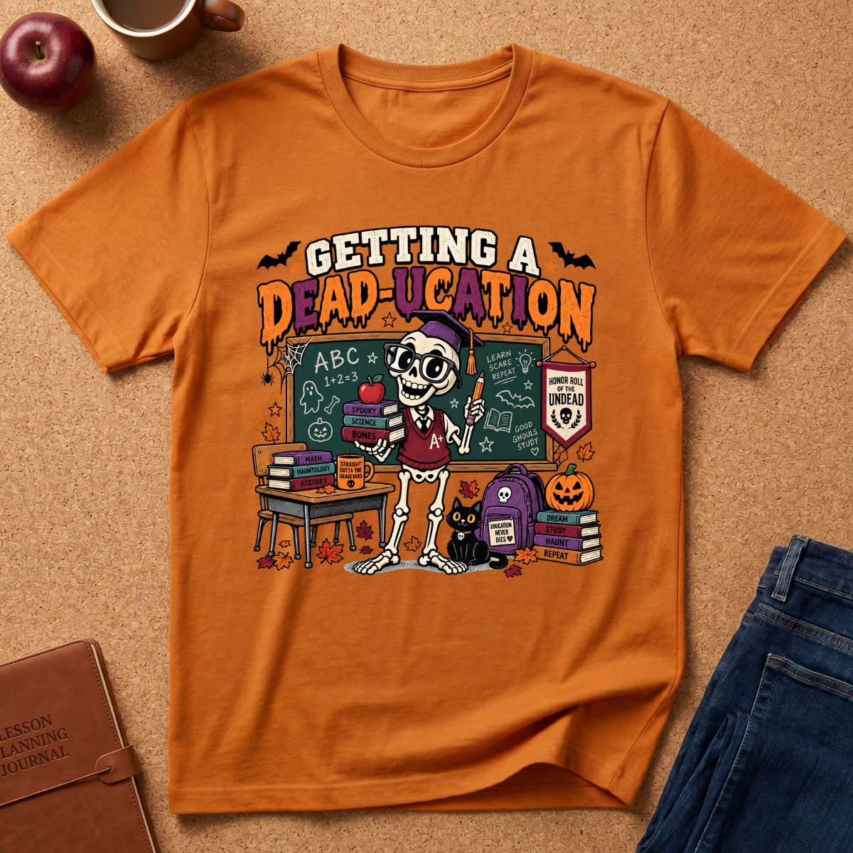 An orange graphic t-shirt laid flat on a cork surface, surrounded by lifestyle props including a brown leather journal, a red apple, a coffee mug, and blue jeans. The t-shirt features a colorful Halloween-themed skeleton teacher graphic with the text 'GETTING A DEAD-UCATION'.