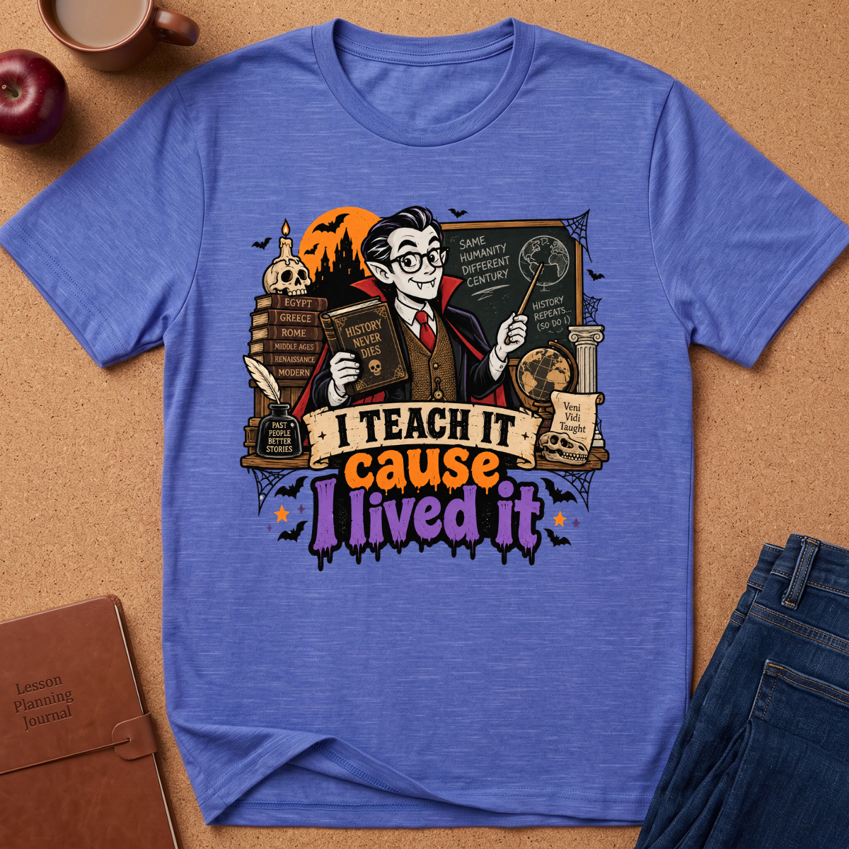 A heathered blue short-sleeve graphic T-shirt laid flat on a cork background, featuring a whimsical illustrated teacher character with books, a wand, and the text 'I TEACH IT cause I lived it.' Surrounding props include an apple, a mug, a notebook, and folded jeans.
