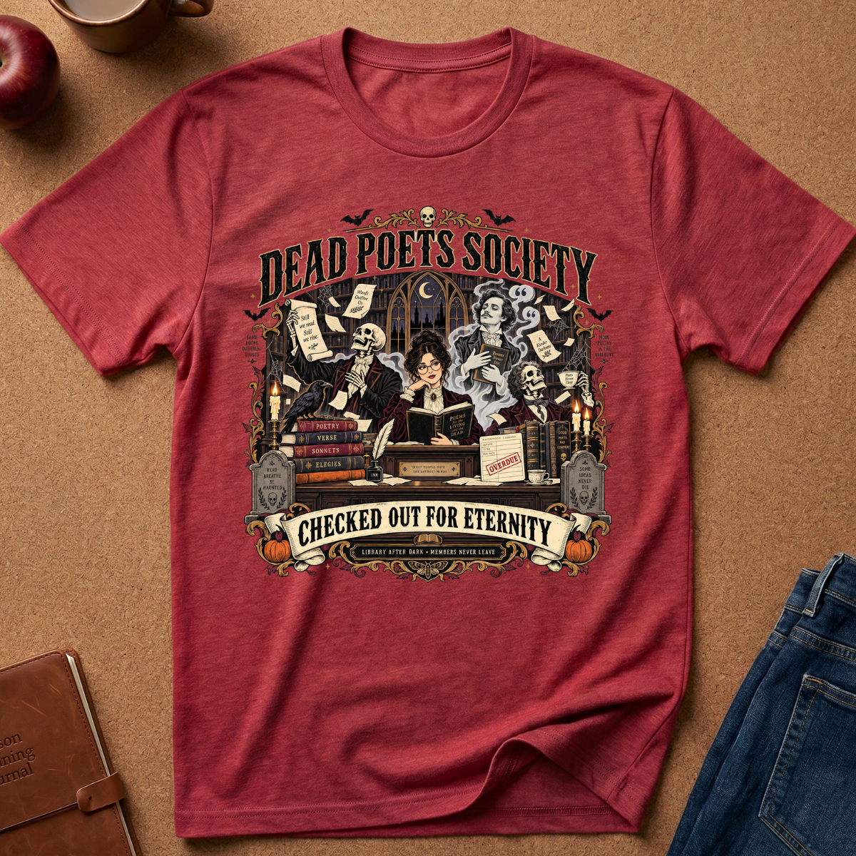 A heathered red graphic t-shirt laid flat on a brown cork background, styled with a notebook, apple, mug, and blue jeans. The shirt features a detailed vintage-style illustration of a witch reading with skeletons and scrolls, with the text 'DEAD POETS SOCIETY' and 'CHECKED OUT FOR ETERNITY'.