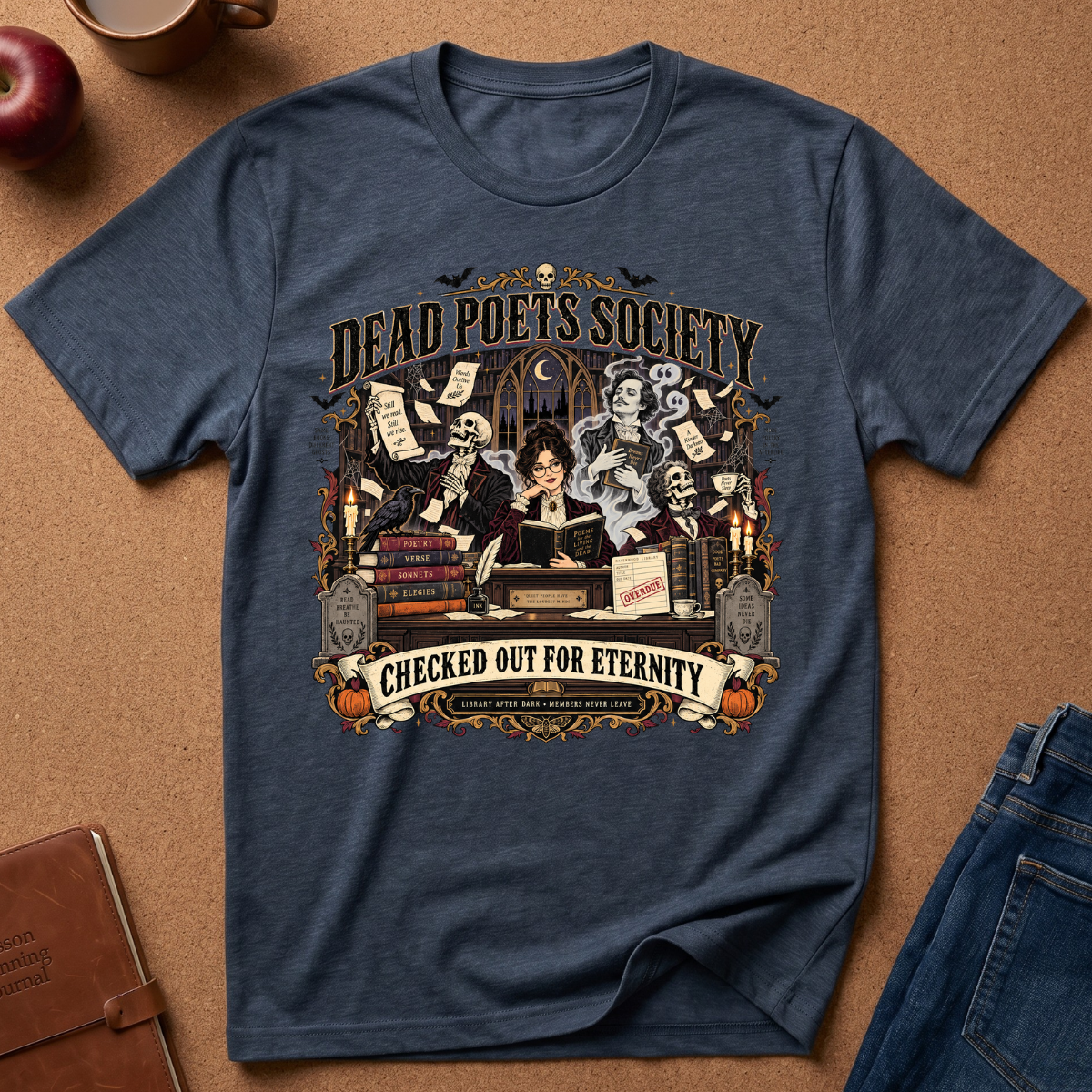 A navy blue graphic t-shirt laid flat on a brown cork background, surrounded by lifestyle props like an apple, a mug, a notebook, and jeans. The shirt features a detailed gothic illustration of a woman surrounded by skeletons, books, and scrolls, with the text 'DEAD POETS SOCIETY' and 'CHECKED OUT FOR ETERNITY'.