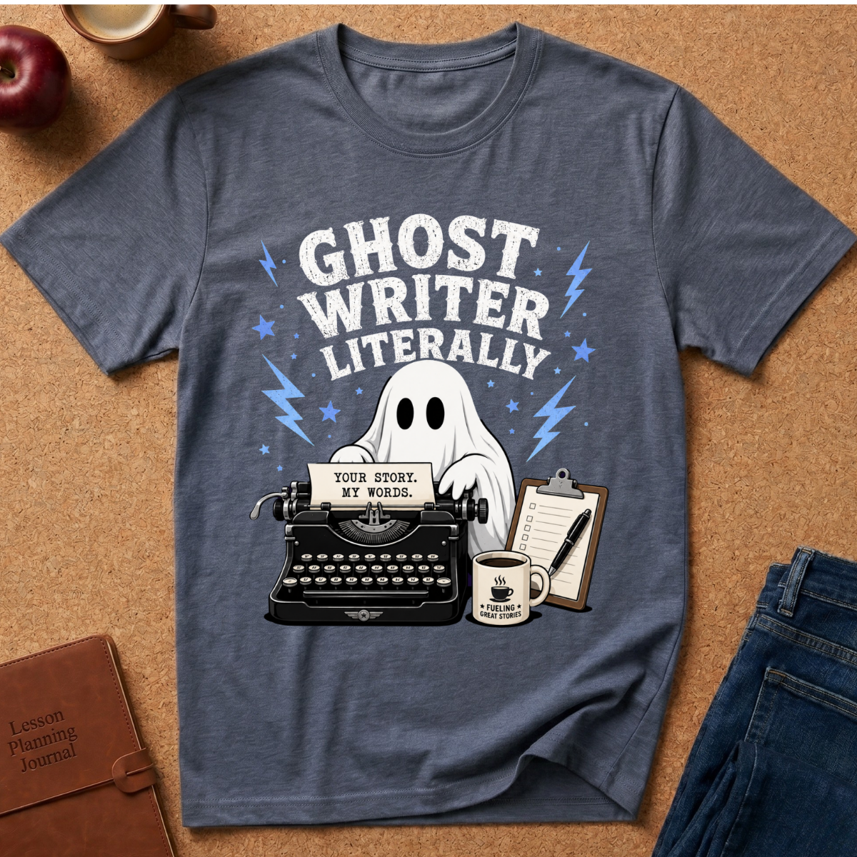 A heathered slate blue graphic t-shirt laid flat on a cork background, featuring a 'Ghost Writer Literally' design with a typewriter, ghost, coffee mug, and notebook.