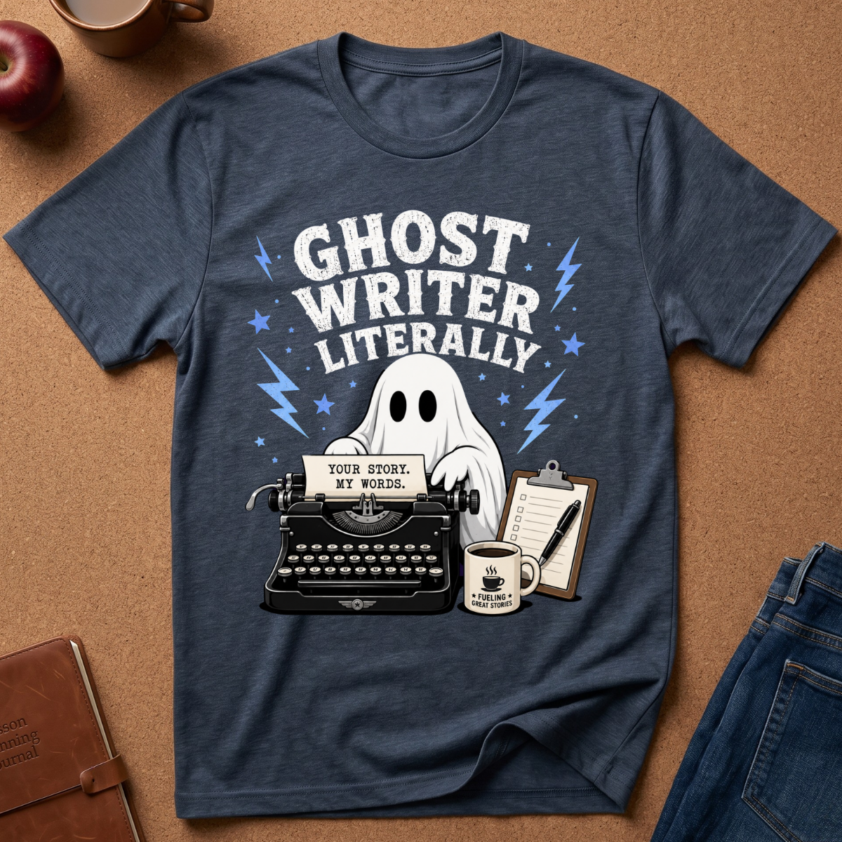 A navy blue graphic t-shirt laid flat on a brown cork background, featuring a white ghost, typewriter, coffee mug, and the text 'GHOST WRITER LITERALLY'.