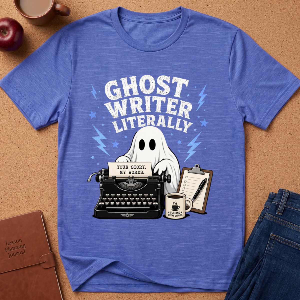 A heathered blue graphic t-shirt laid flat on a cork background, featuring a ghost writer illustration with a typewriter, notebook, coffee mug, and the text 'GHOST WRITER LITERALLY'.