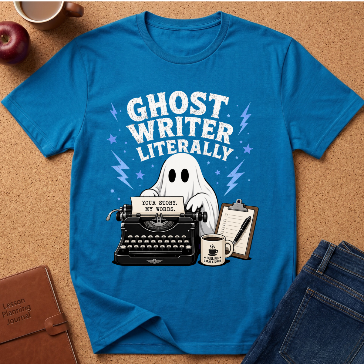 A bright blue graphic T-shirt laid flat on a cork surface, surrounded by styled props including a coffee mug, apple, journal, and jeans. The shirt features a ghostwriter-themed print with the words “GHOST WRITER LITERALLY,” a white ghost, typewriter, notebook, clipboard, and coffee mug.