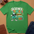 A green short-sleeve graphic T-shirt laid flat on a cork surface, with a cartoon beaker character and the words 'SCIENCE TEACHER MAKING REACTIONS HAPPEN EVERY DAY' printed on the front. Blue jeans, a brown notebook, and a mug are partially visible around it as styling props.