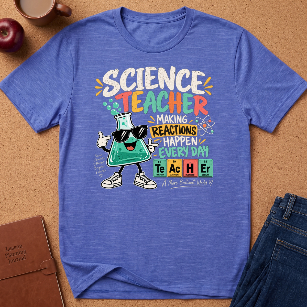 A heathered blue graphic T-shirt laid flat on a cork surface, surrounded by styled props including an apple, a mug, a notebook, and jeans. The shirt features a colorful 'Science Teacher' design with a cartoon beaker wearing sunglasses and periodic-table-style element blocks spelling TeAcHEr.