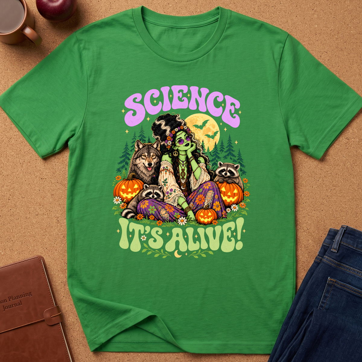 A green short-sleeve graphic T-shirt laid flat on a cork surface, surrounded by a coffee mug, apple, notebook, and jeans. The shirt features a colorful Halloween-themed illustration with bats, pumpkins, raccoons, a cat, and a woman, plus the text 'SCIENCE IT'S ALIVE!'.