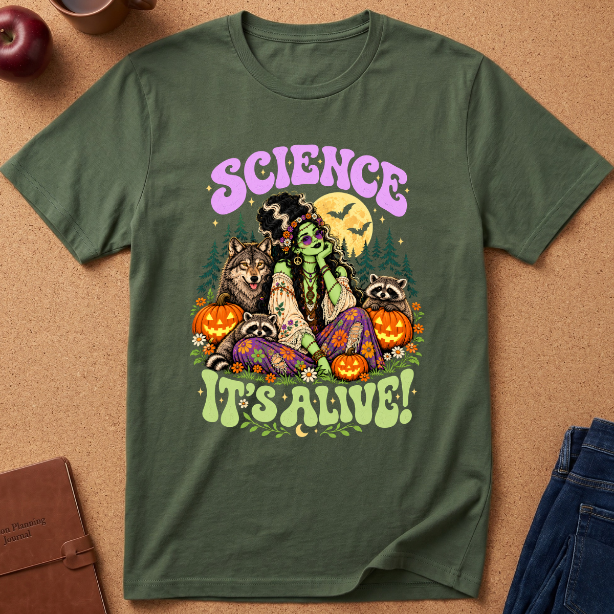 An olive green graphic t-shirt laid flat on a cork background, featuring a colorful Halloween-themed illustration of a green-skinned woman, a wolf, raccoons, pumpkins, bats, and the text 'SCIENCE IT'S ALIVE!'.