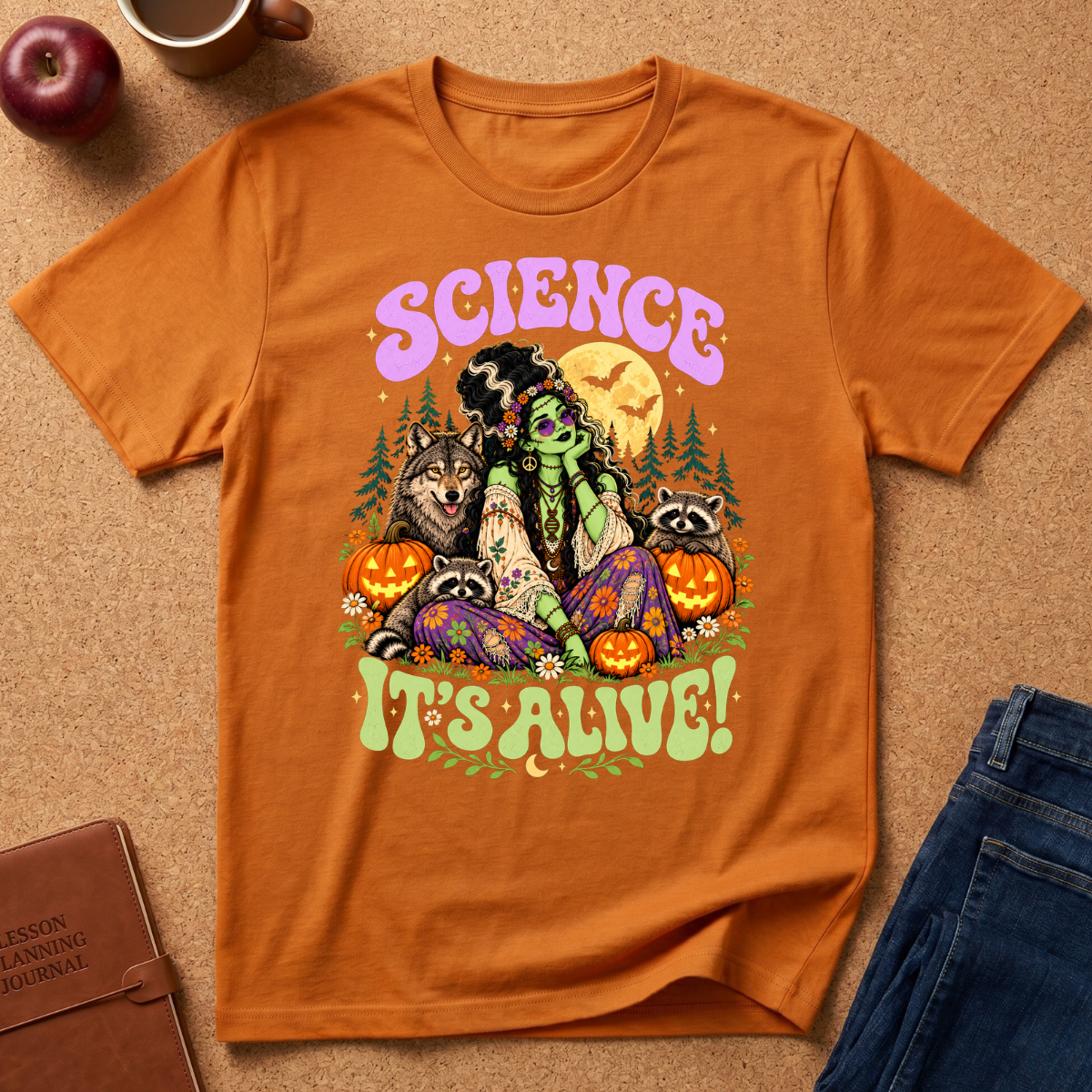 An orange graphic t-shirt laid flat on a cork background, featuring a colorful illustration of a witch surrounded by raccoons, pumpkins, and the text 'SCIENCE IT'S ALIVE!'. Styled flat-lay props include an apple, a coffee mug, a journal, and folded blue jeans.