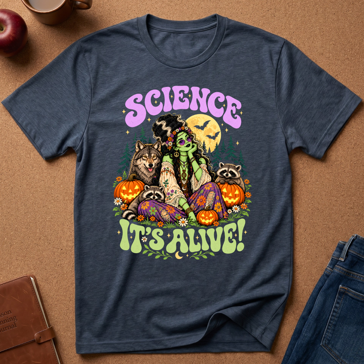 A navy blue graphic t-shirt laid flat on a brown cork background, featuring a colorful Halloween-themed illustration with the text 'SCIENCE IT'S ALIVE!'. The design includes a green monster, raccoons, a wolf, pumpkins, and bats.