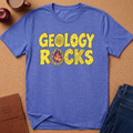 A heathered blue short-sleeve t-shirt laid flat on a cork background, featuring the words 'GEOLOGY ROCKS' in yellow block letters with a geode illustration inside the letter O. Styled flat-lay props include a brown journal, an apple, a coffee mug, and blue jeans.
