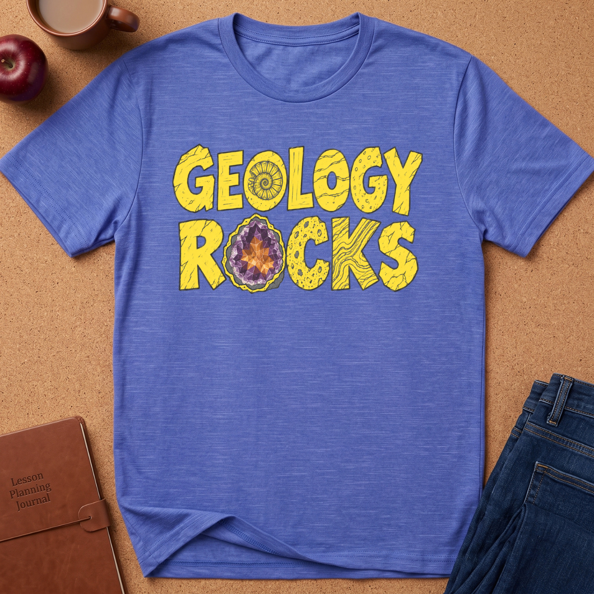 A heathered blue short-sleeve t-shirt laid flat on a cork background, featuring the words 'GEOLOGY ROCKS' in yellow block letters with a geode illustration inside the letter O. Styled flat-lay props include a brown journal, an apple, a coffee mug, and blue jeans.