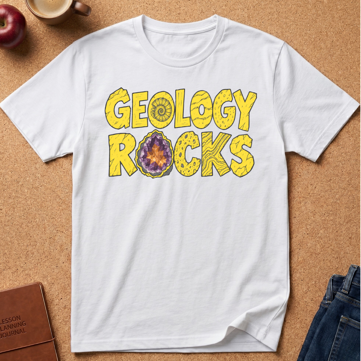 A white short-sleeve t-shirt laid flat on a cork background, featuring a yellow 'GEOLOGY ROCKS' graphic with a geode design inside the letter O. Styled flat-lay props include an apple, a coffee cup, a journal, and jeans.