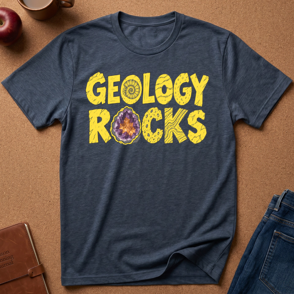 A heather navy blue t-shirt with a yellow 'GEOLOGY ROCKS' graphic printed on the chest, styled in a flat-lay on a brown cork background with an apple, notebook, and jeans.