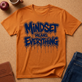 An orange crewneck t-shirt with a dark blue dripping graffiti-style graphic that reads 'Mindset Means Everything'. The shirt is presented in a flat-lay arrangement on a cork background, surrounded by lifestyle props including a brown leather journal, a red apple, a coffee mug, and a pair of blue jeans.