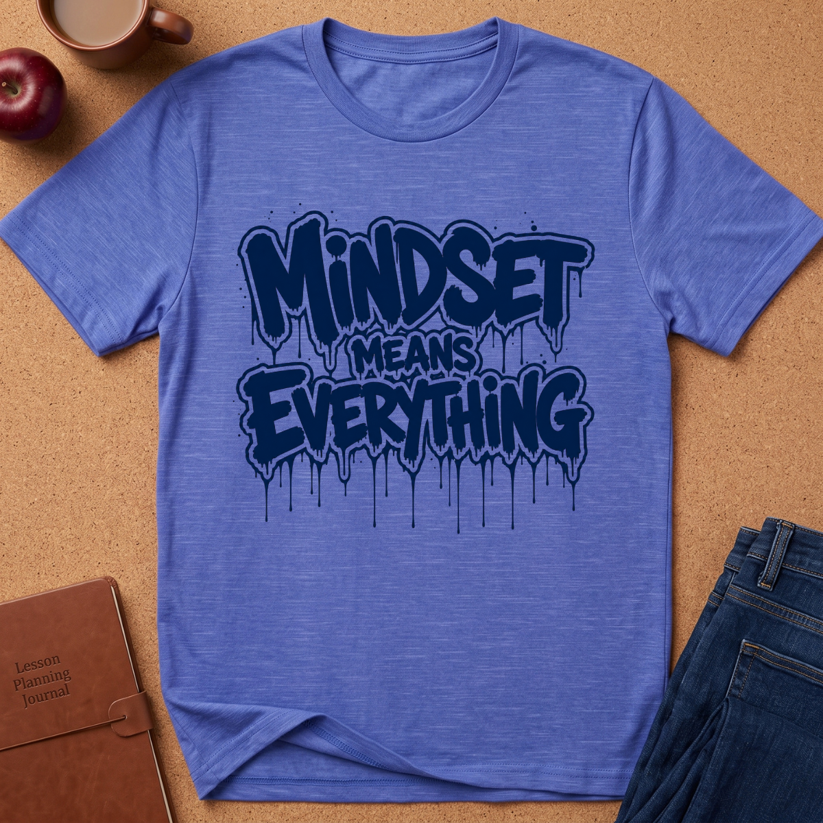 A heathered blue crewneck t-shirt with a dark blue dripping graphic that reads 'Mindset Means Everything' in a graffiti-style font. The shirt is laid flat on a cork background with lifestyle props including a brown leather journal, a red apple, a coffee mug, and blue jeans.