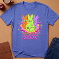 A heathered blue short-sleeve t-shirt laid flat on a cork surface, featuring a colorful graphic of three stitched bunny characters in orange, green, and purple with the text 'Chillin with my CREEPS'. A brown notebook, apple, mug, and jeans are partially visible around the shirt as styled props.