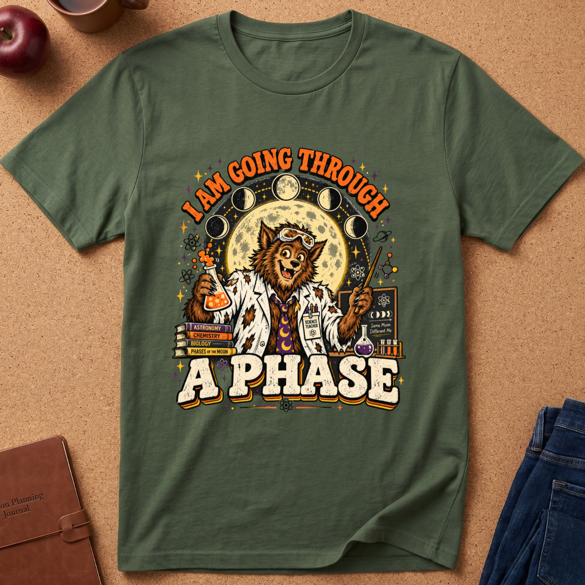 An olive green graphic t-shirt laid flat on a cork background, featuring a detailed illustration of a werewolf in a lab coat with the text 'I AM GOING THROUGH A PHASE'.