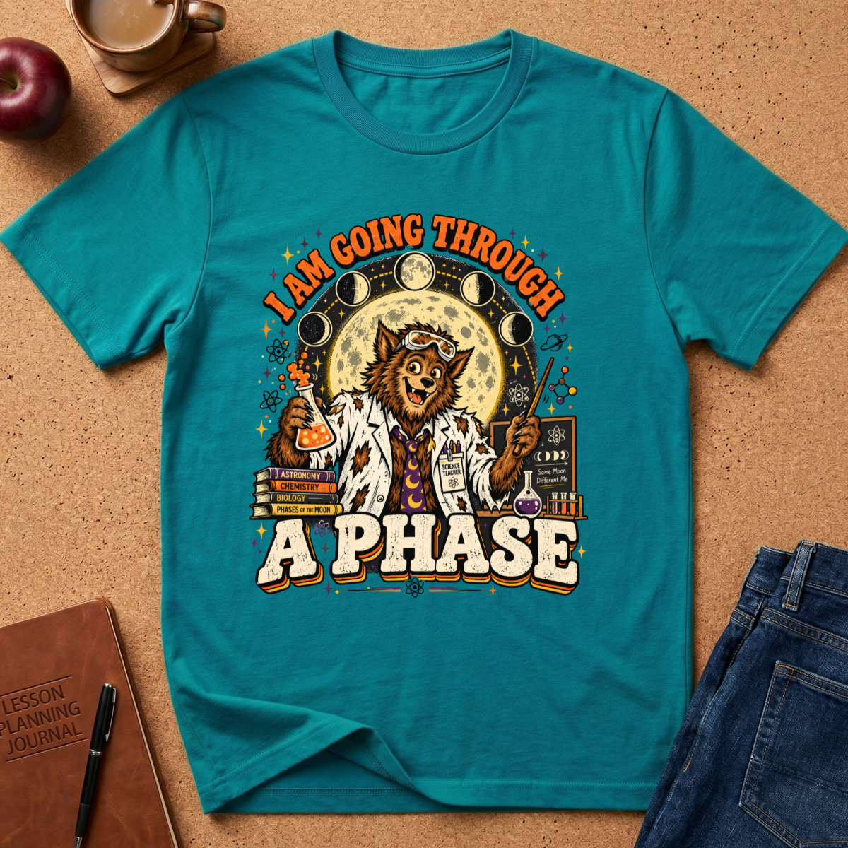 A teal graphic t-shirt laid flat on a cork surface with styled props including an apple, coffee cup, notebook, and jeans. The shirt features a cartoon cat in a lab coat with moon phases, beakers, and the text 'I AM GOING THROUGH A PHASE'.