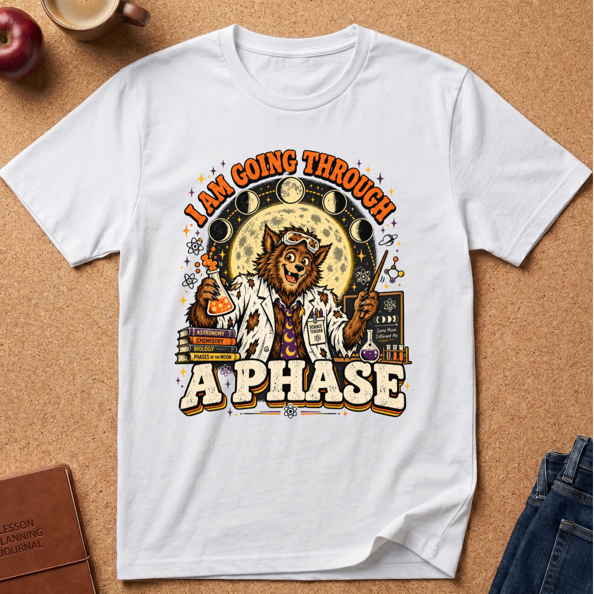 A white short-sleeve graphic T-shirt laid flat on a cork background, featuring a cartoon cat in a lab coat with the text 'I AM GOING THROUGH A PHASE' and moon-phase and chemistry-themed artwork. Surrounding props include an apple, a coffee cup, a journal, and folded jeans.