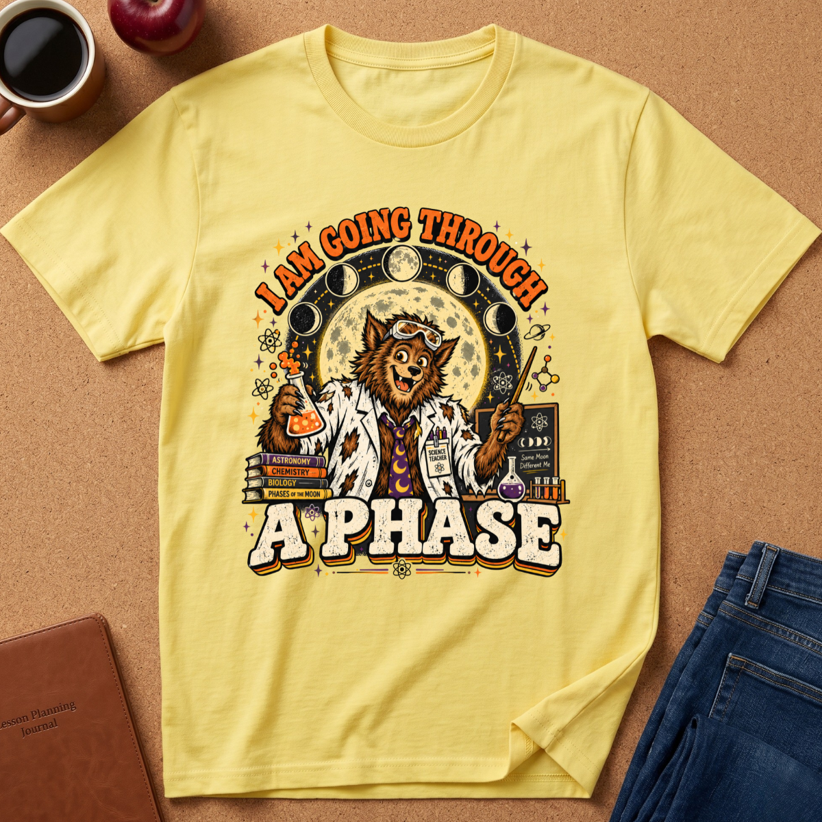 A pale yellow graphic T-shirt laid flat on a cork surface with styled props including a coffee mug, apple, notebook, and jeans. The shirt features a large illustrated wolf in a lab coat surrounded by moon phases, books, and chemicals, with the text 'I AM GOING THROUGH A PHASE'.