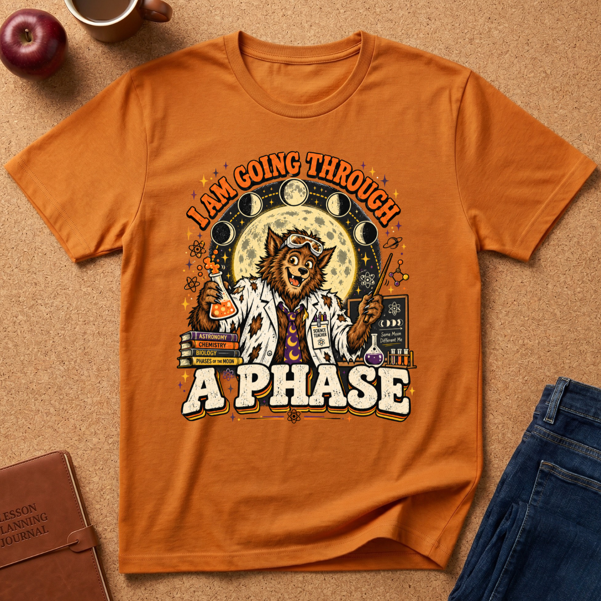 An orange short-sleeve graphic T-shirt laid flat on a cork surface, surrounded by lifestyle props including an apple, a mug, a journal, and folded blue jeans. The shirt features a large front print of a labrador retriever in a white lab coat surrounded by moon phases, books, and chemicals, with the text 'I AM GOING THROUGH A PHASE'.