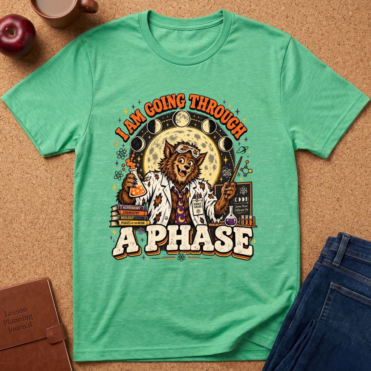 A green graphic t-shirt laid flat on a cork background, featuring a detailed illustration of a cat dressed as a scientist with the text 'I AM GOING THROUGH A PHASE'. The shirt is styled with a brown leather journal, an apple, a coffee mug, and blue jeans.
