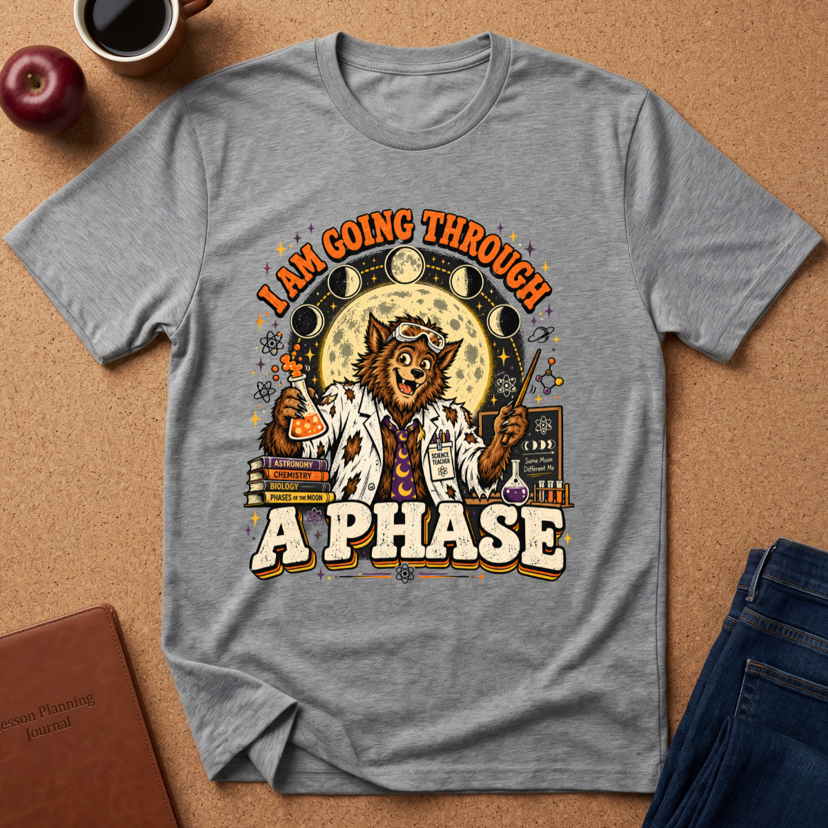 A heather gray graphic t-shirt laid flat on a cork background, featuring a whimsical design of a cat dressed as a scientist with the text 'I AM GOING THROUGH A PHASE'. The shirt is styled with a coffee mug, apple, notebook, and jeans nearby.
