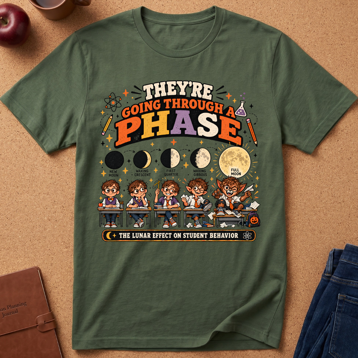 An olive green graphic t-shirt laid flat on a cork background, featuring a moon phase and cartoon student illustration with the text 'THEY'RE GOING THROUGH A PHASE' and 'THE LUNAR EFFECT OR STUDENT BERVOR'.
