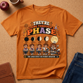 An orange graphic T-shirt laid flat on a cork surface with styled props including an apple, mug, notebook, and jeans. The shirt features a large moon-phase-themed school behavior print with cartoon students and text reading 'THEY'RE GOING THROUGH A PHASE' and 'THE LUNAR EFFECT ON STUDENT BEHAVIOR.