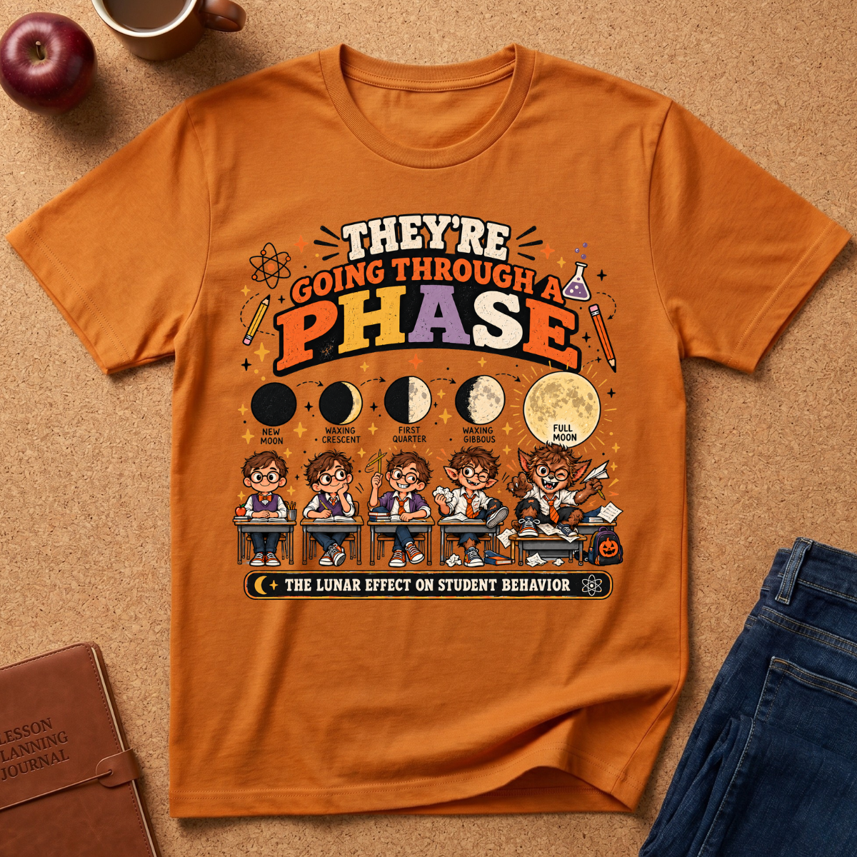 An orange graphic T-shirt laid flat on a cork surface with styled props including an apple, mug, notebook, and jeans. The shirt features a large moon-phase-themed school behavior print with cartoon students and text reading 'THEY'RE GOING THROUGH A PHASE' and 'THE LUNAR EFFECT ON STUDENT BEHAVIOR.