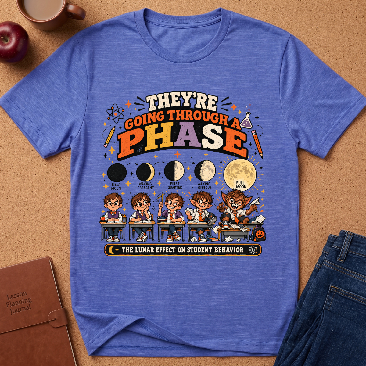 A heathered blue graphic t-shirt laid flat on a brown cork background, surrounded by lifestyle props including a coffee cup, apple, notebook, and jeans. The shirt features a detailed graphic about the lunar cycle and student behavior.