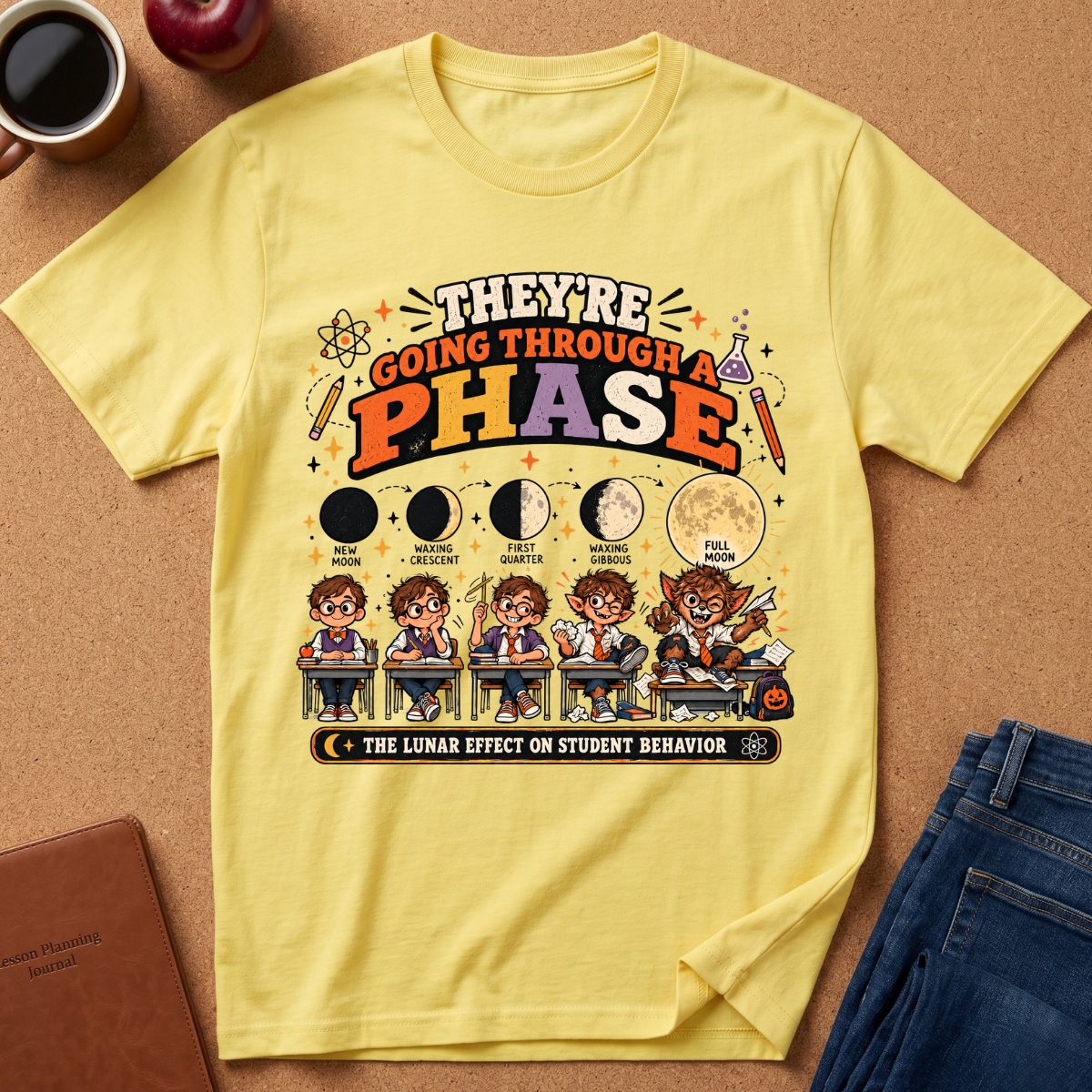 A yellow graphic t-shirt laid flat on a brown corkboard background, surrounded by lifestyle props including a coffee mug, an apple, a notebook, and blue jeans. The t-shirt features a colorful cartoon graphic of students and moon phases with the text 'THEY'RE GOING THROUGH A PHASE - THE LUNAR EFFECT ON STUDENT BEHAVIOR'.