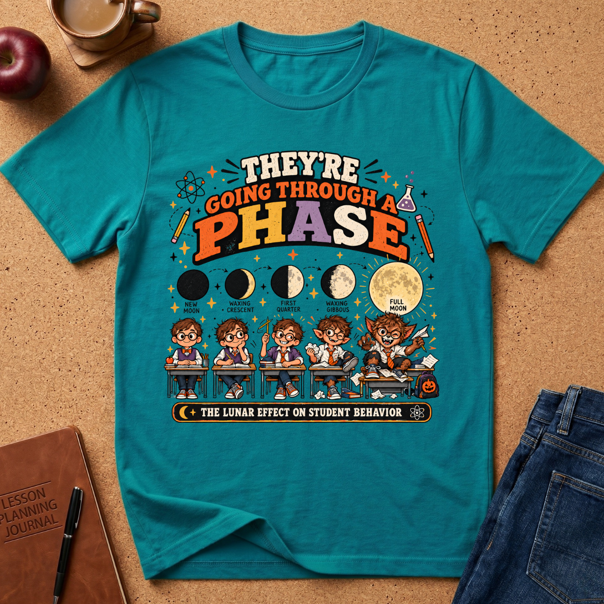 A teal graphic t-shirt laid flat on a cork surface with styled props including an apple, coffee cup, notebook, and jeans. The shirt features a colorful educational moon-phase design with cartoon students and the text 'THEY'RE GOING THROUGH A PHASE THE LUNAR EFFECT ON STUDENT BEHAVIOR'.