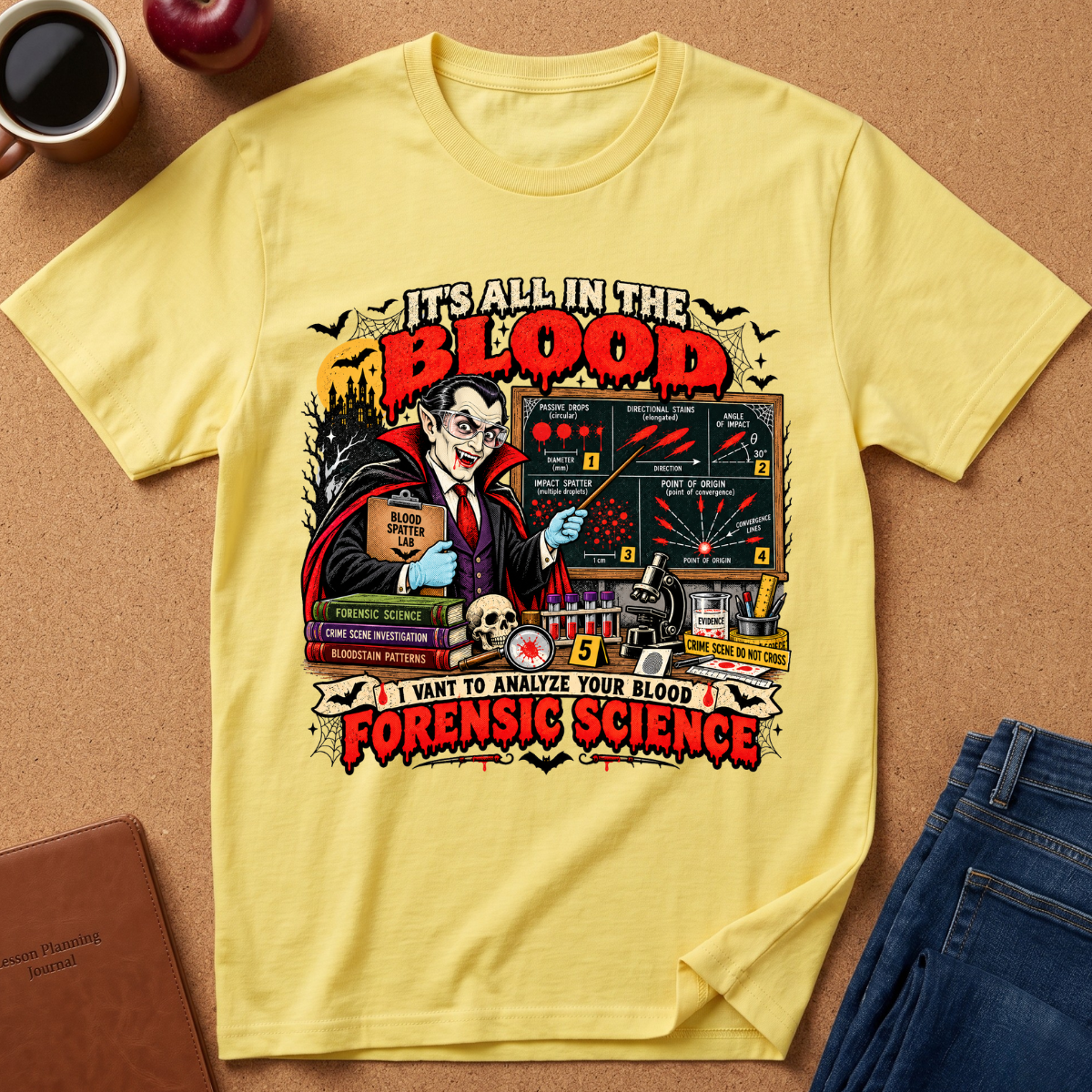 A yellow graphic t-shirt featuring a vintage-style forensics and vampire design, laid flat on a cork background with lifestyle props including jeans, a coffee mug, and a notebook.