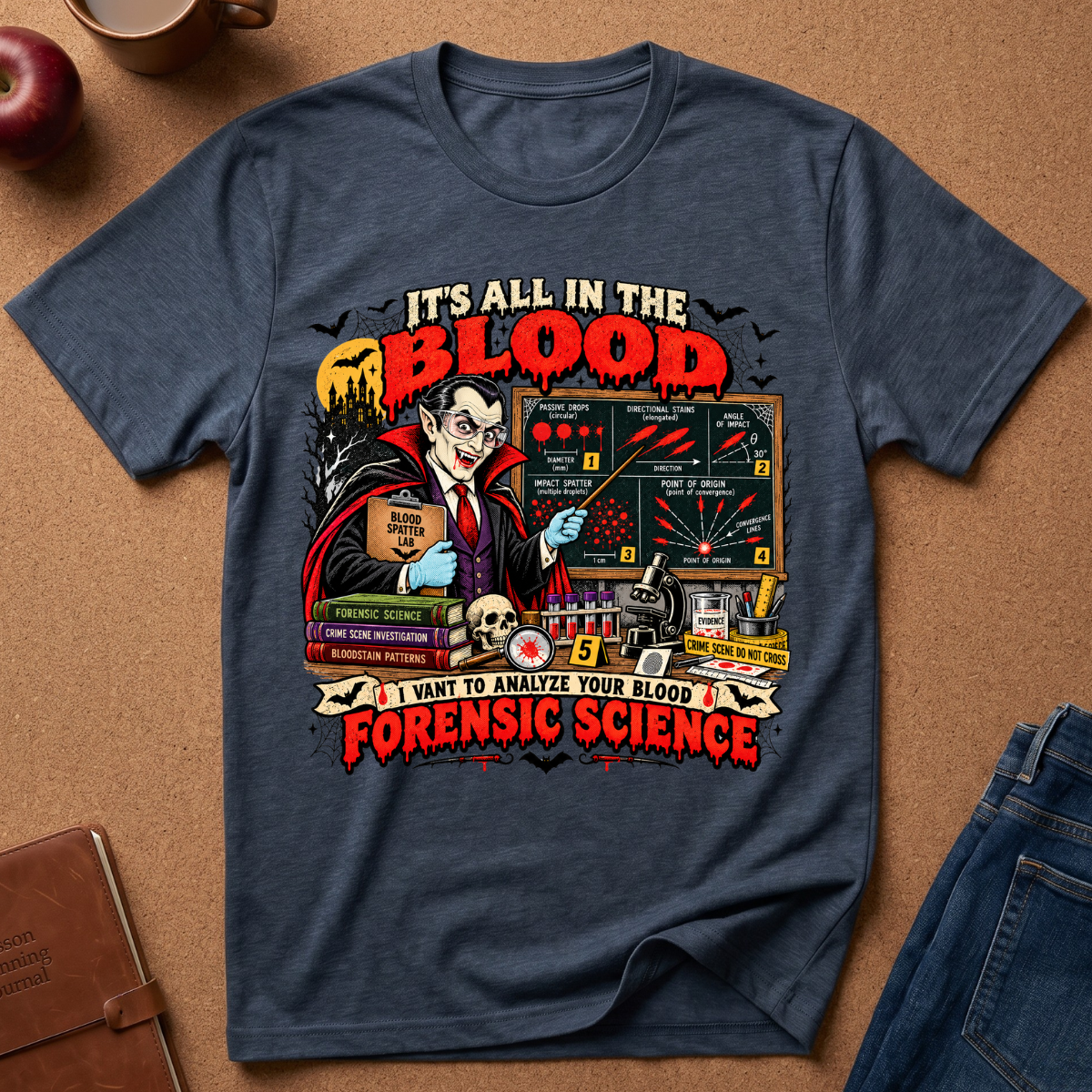 A navy blue graphic t-shirt laid flat on a brown cork board background, styled with a leather journal, an apple, and blue jeans. The t-shirt features a detailed graphic of a vampire scientist with a blood test kit, the text 'IT'S ALL IN THE BLOOD' and 'I WANT TO ANALYZE YOUR BLOOD FORENSIC SCIENCE'.