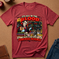 A red graphic t-shirt laid flat on a brown textured surface, surrounded by styled props including an apple, a mug, a notebook, and blue jeans. The shirt features a detailed vintage-style illustration of a vampire scientist with blood tests, bats, and the text 'IT'S ALL IN THE BLOOD' and 'I WANT TO ANALYZE YOUR BLOOD FORENSIC SCIENCE'.