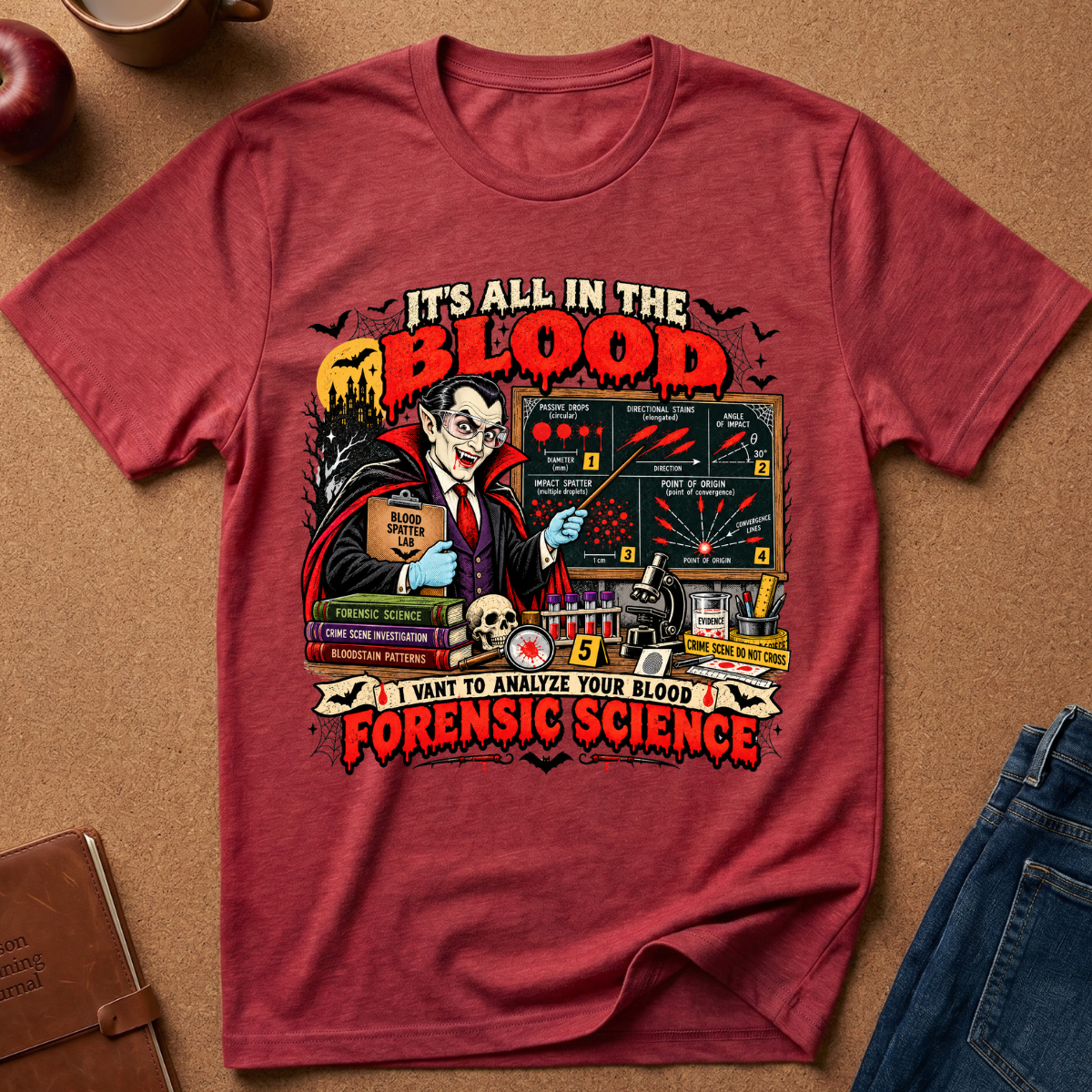 A red graphic t-shirt laid flat on a brown textured surface, surrounded by styled props including an apple, a mug, a notebook, and blue jeans. The shirt features a detailed vintage-style illustration of a vampire scientist with blood tests, bats, and the text 'IT'S ALL IN THE BLOOD' and 'I WANT TO ANALYZE YOUR BLOOD FORENSIC SCIENCE'.