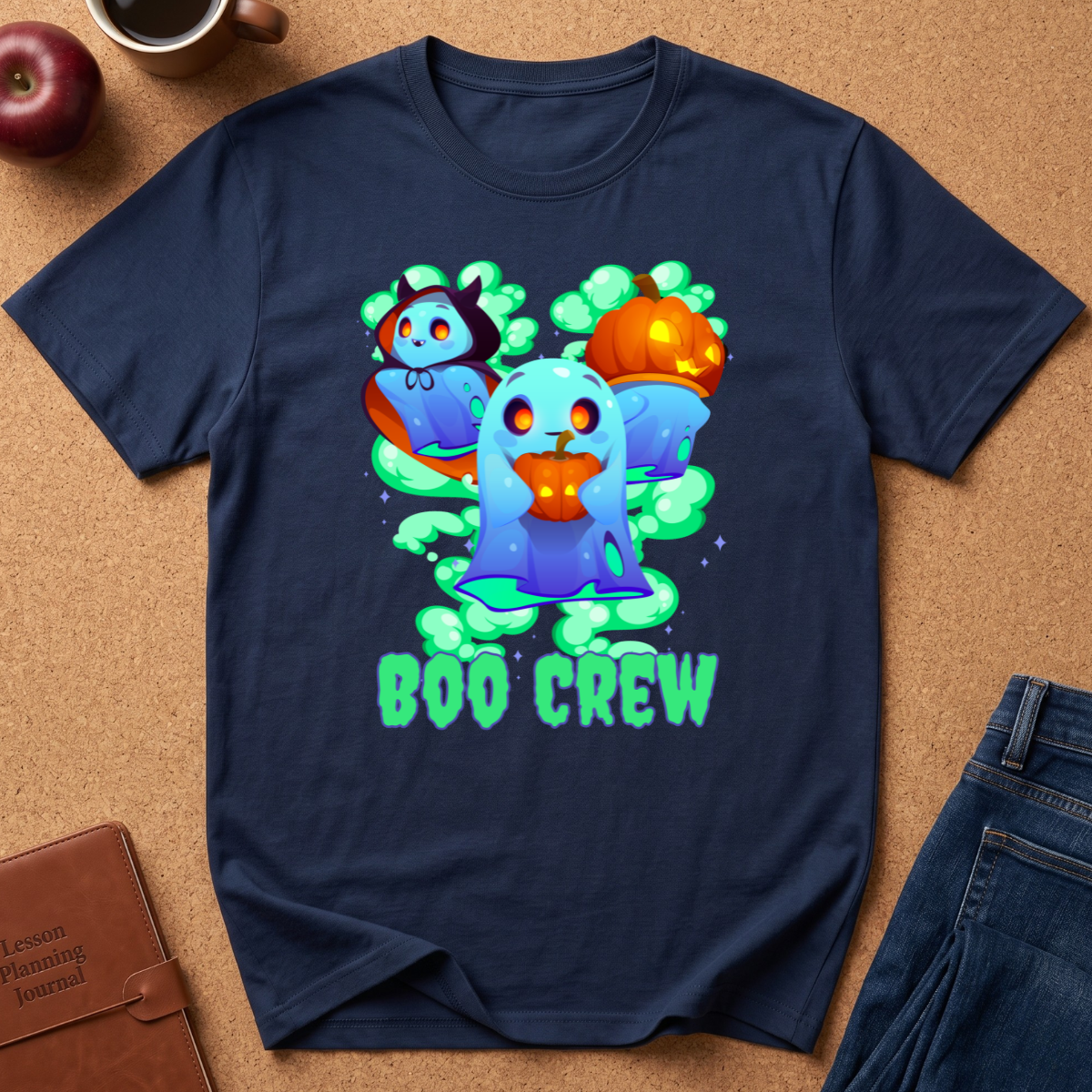 A navy blue short-sleeve t-shirt laid flat on a brown cork background, featuring a colorful graphic of three cartoon ghosts with Halloween-themed accessories and the words 'BOO CREW' in bright green. Partially visible props around the shirt include an apple, a coffee cup, a brown journal, and blue jeans.