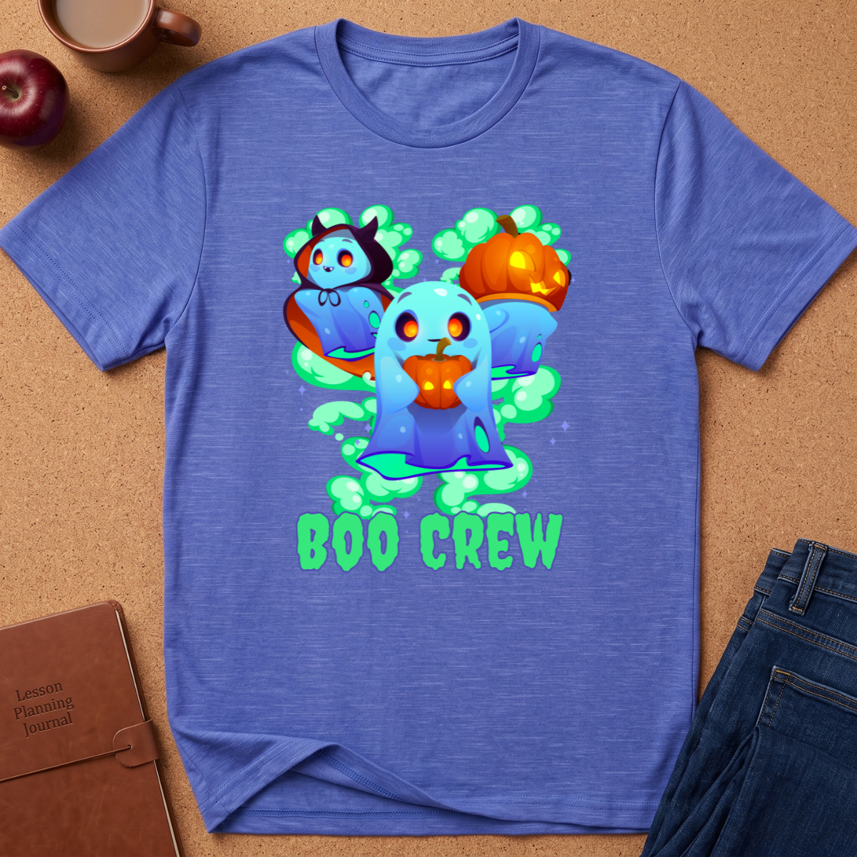 A heathered blue short-sleeve t-shirt laid flat on a brown cork background, featuring a colorful graphic of three cartoon ghosts with pumpkins and the text 'BOO CREW'. Surrounding props include an apple, a coffee mug, a notebook, and jeans.