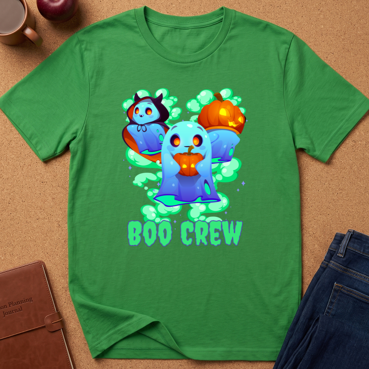 A green short-sleeve t-shirt laid flat on a cork surface, featuring a colorful graphic of three cartoon ghosts with pumpkins and the text 'BOO CREW'. Partial props are visible at the edges, including a brown leather journal, an apple, a mug, and blue jeans.
