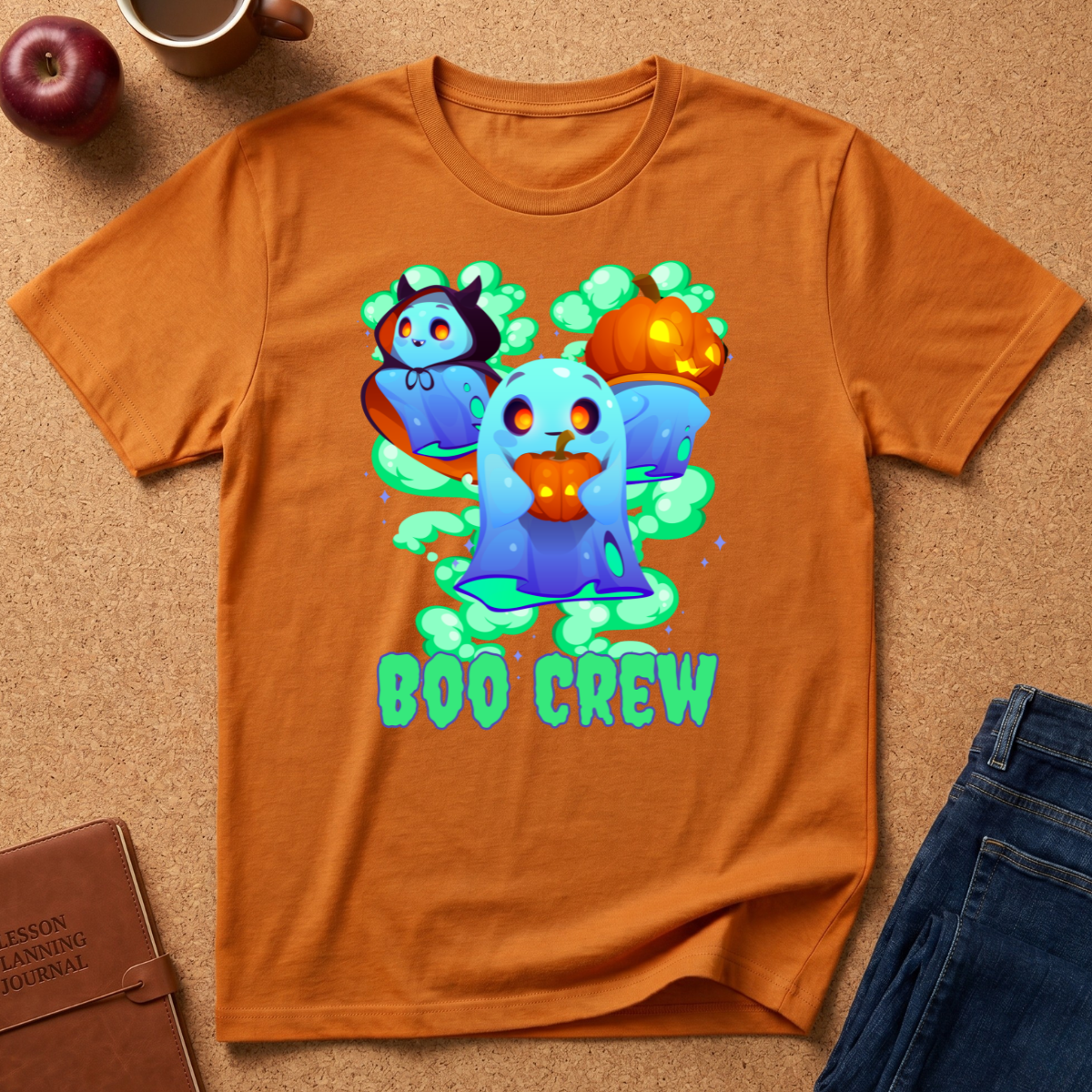 An orange crewneck t-shirt with a colorful cartoon graphic of a blue ghost, a vampire, a jack-o'-lantern, and the text 'BOO CREW' in green, styled in a flat-lay on a cork background with an apple, notebook, and jeans.