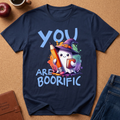 A navy blue graphic t-shirt laid flat on a cork background, featuring a cute cartoon ghost in a witch hat holding a colorful pencil and holding up a sign that says 'YOU ARE BOORIFIC'.
