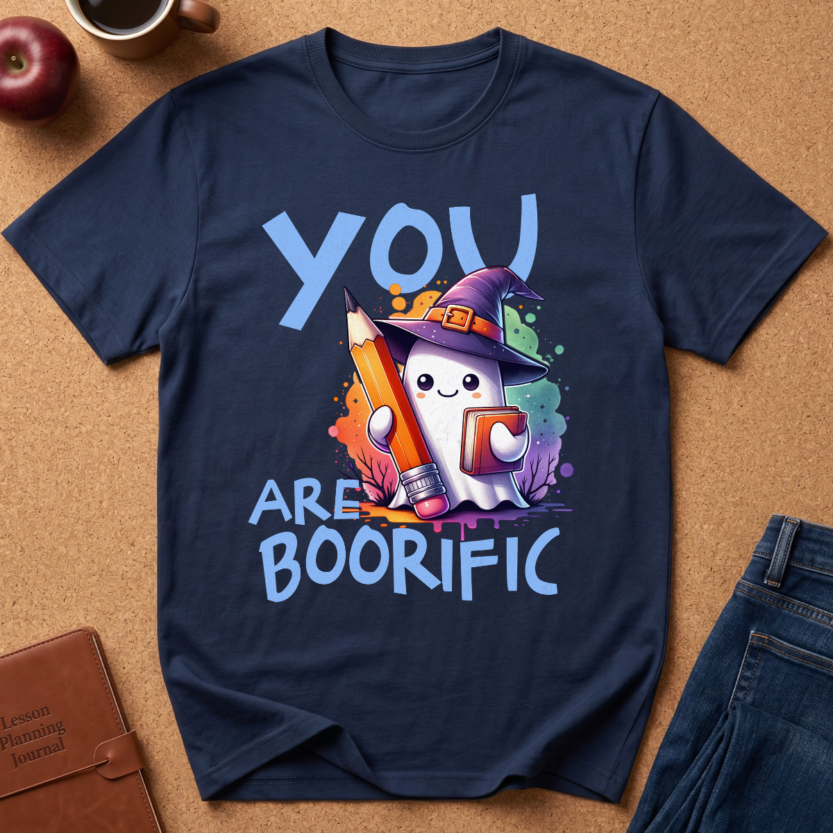 A navy blue graphic t-shirt laid flat on a cork background, featuring a cute cartoon ghost in a witch hat holding a colorful pencil and holding up a sign that says 'YOU ARE BOORIFIC'.