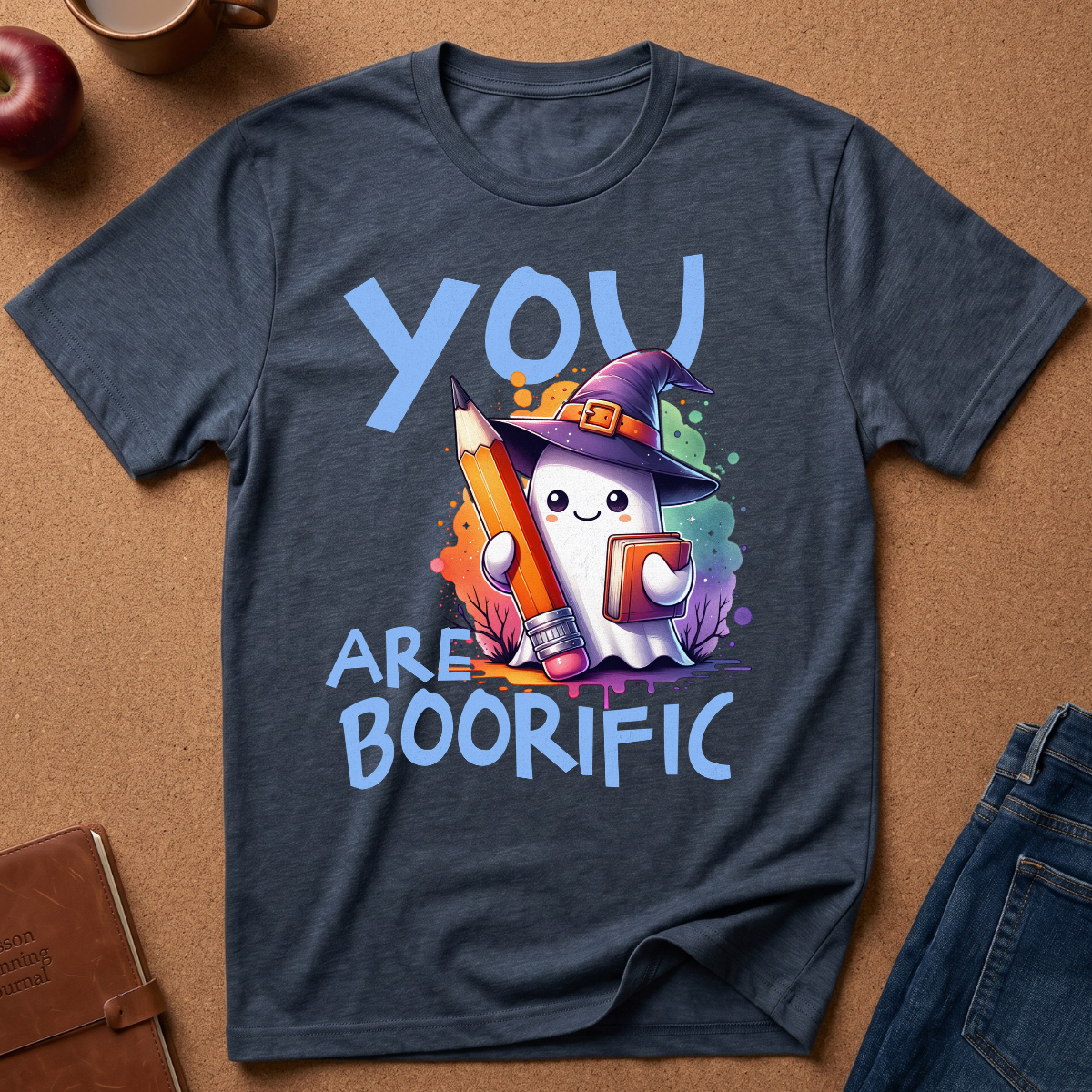 A dark heather blue graphic t-shirt laid flat on a brown cork background, featuring a cute cartoon ghost in a witch hat holding a pencil and the text 'YOU ARE BOORIFIC'.