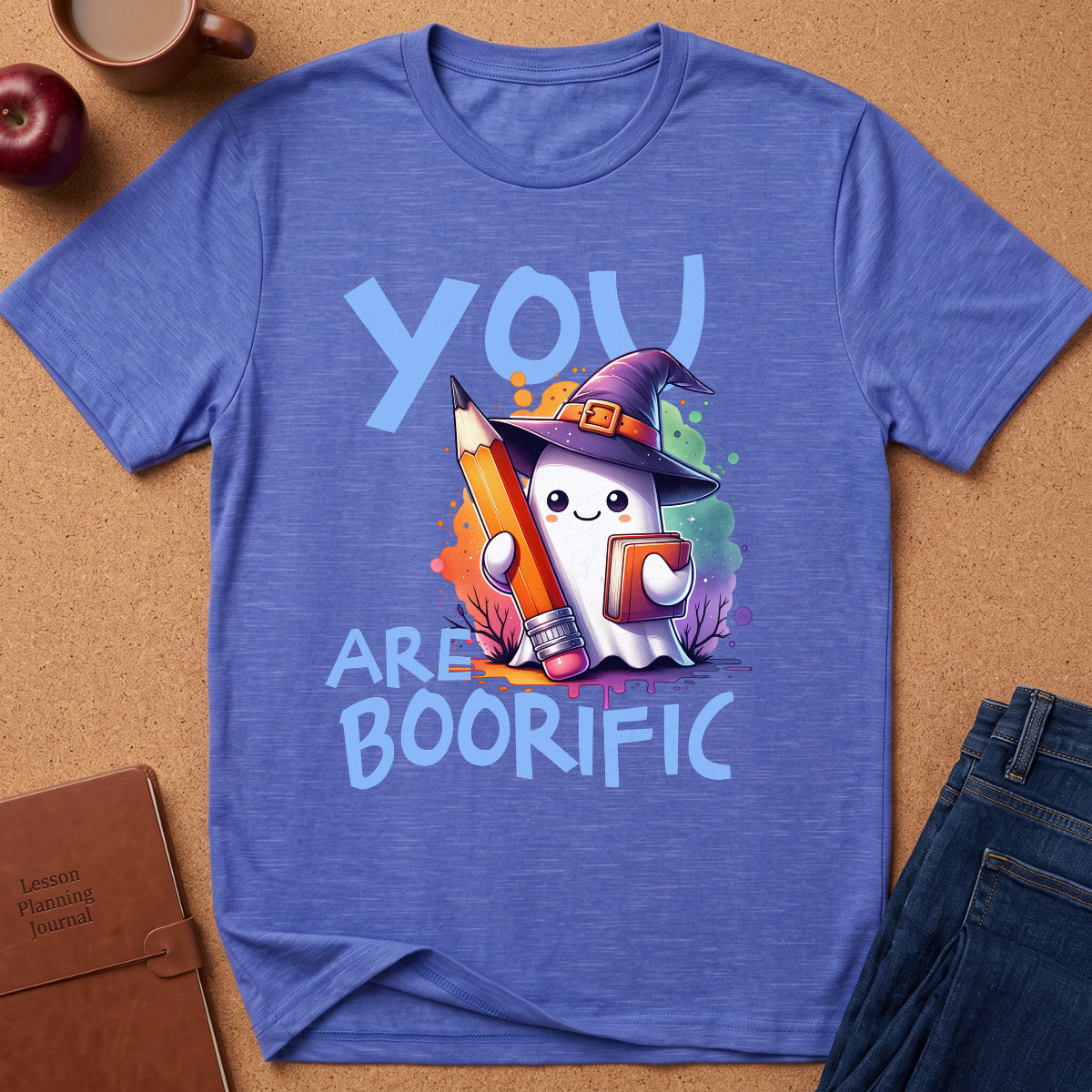 A heathered blue graphic t-shirt laid flat on a brown cork background, surrounded by lifestyle props including a coffee mug, an apple, a notebook, and jeans. The t-shirt features a cute cartoon ghost wearing a witch hat holding a large orange pencil, with the text 'YOU ARE BOORIFIC' in light blue.