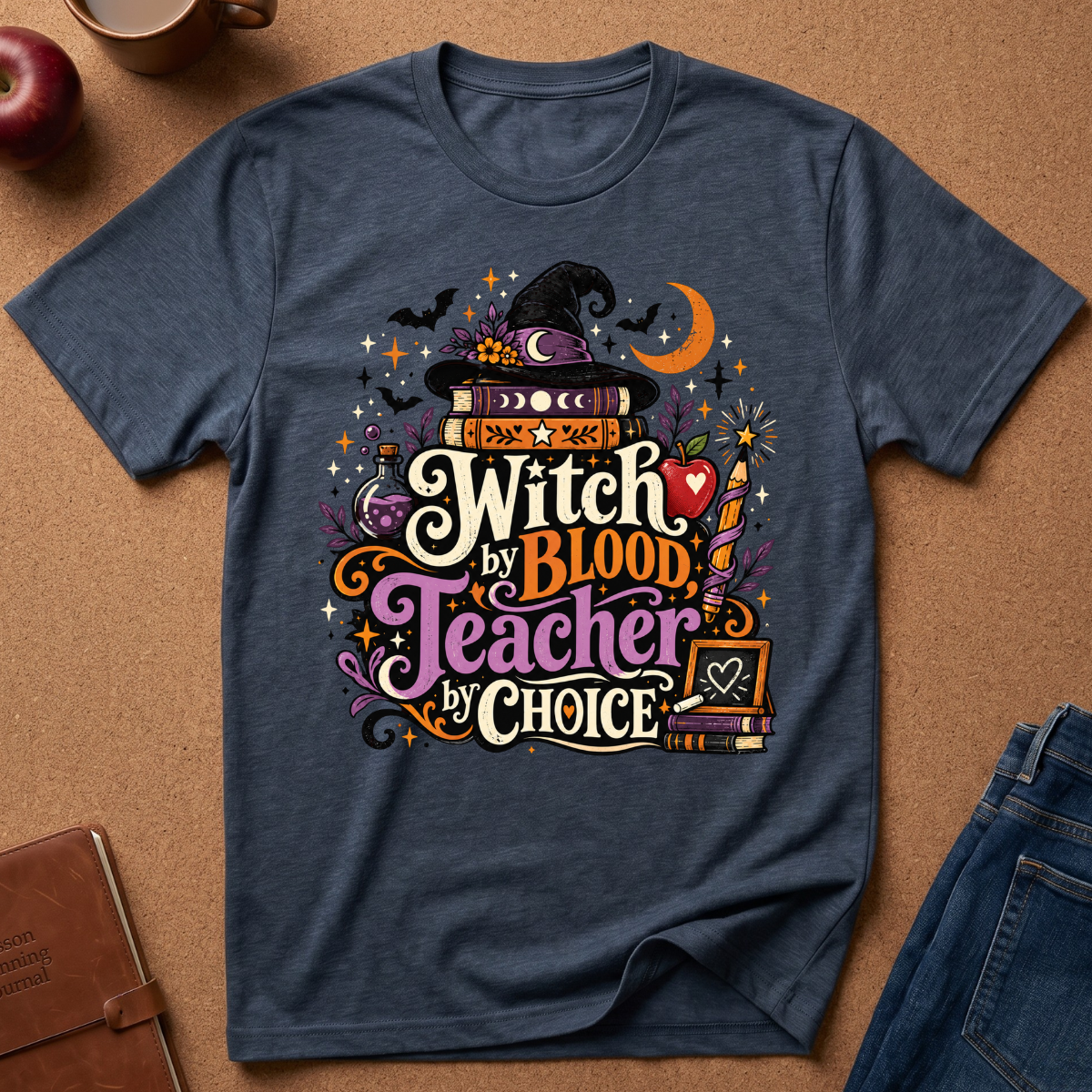 A navy blue graphic t-shirt laid flat on a brown cork background, surrounded by lifestyle props like an apple, a mug, a notebook, and jeans. The shirt features a colorful Halloween-themed graphic with the text 'Witch by Blood Teacher by Choice' and illustrations of a witch hat, potion bottle, bats, and a crescent moon.