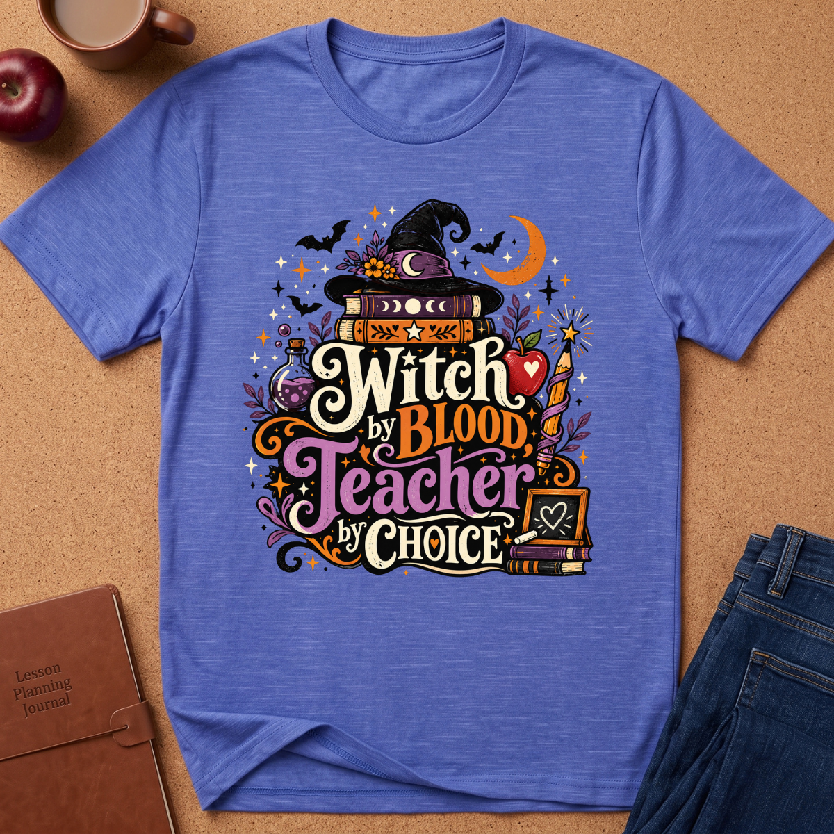 A heathered blue short-sleeve t-shirt laid flat on a cork background, featuring a colorful Halloween-themed graphic that reads 'Witch by Blood Teacher by Choice' with a witch hat, books, bats, stars, and a crescent moon. Styled flat-lay props include a brown journal, an apple, a mug, and blue jeans.