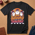 A black short-sleeve graphic T-shirt laid flat on a cork surface, featuring three cartoon ghosts wearing graduation caps and the phrase 'One Spooky Teacher' in orange, pink, and purple lettering. Styled flat-lay props include an apple, a mug, a journal, and jeans around the shirt.
