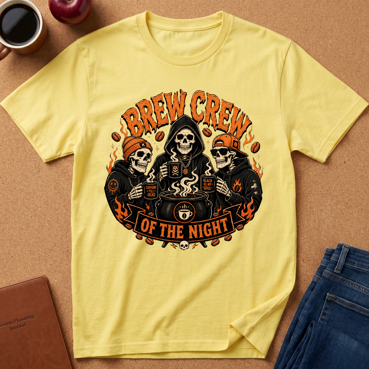 A pale yellow graphic T-shirt laid flat on a cork surface, surrounded by coffee cups, an apple, a notebook, and folded blue jeans. The shirt features a large front print of three skeleton characters in black hoodies with orange flames and the text 'BREW CREW OF THE NIGHT.
